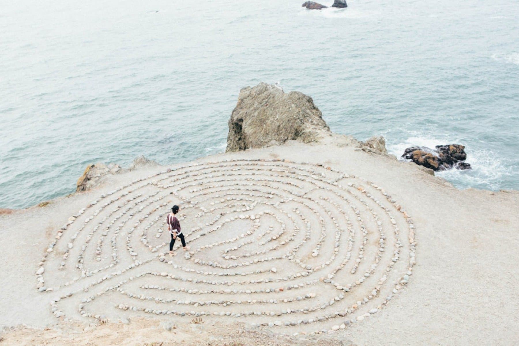 A woman walking a coastal stone labyrinth at the edge of a cliff in soft daylight.